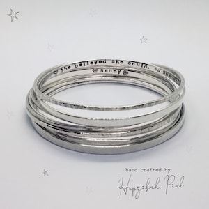 May include: A set of silver bangles with a hammered finish. The bangles are stacked on top of each other and have the inscription "She believed she could, so she did. Nanny" on one of the bangles. The bangles are hand crafted by Hepzibah Pink.