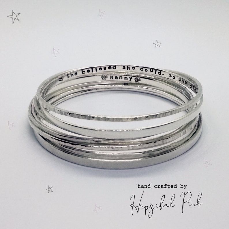 May include: A set of silver bangles with a hammered finish. The bangles are stacked on top of each other and have the inscription "She believed she could, so she did. Nanny" on one of the bangles. The bangles are hand crafted by Hepzibah Pink.