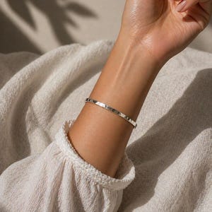 Peut inclure: Un bracelet argenté à texture martelée est porté au poignet. Le bracelet est de conception simple. La personne porte une chemise blanche à manches longues avec un col à volants. L'arrière-plan est de couleur neutre.