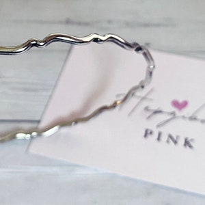 May include: A silver wavy bangle bracelet. The bracelet is on a white surface with a pink heart and the text "Hashtag Pink" on a white card.