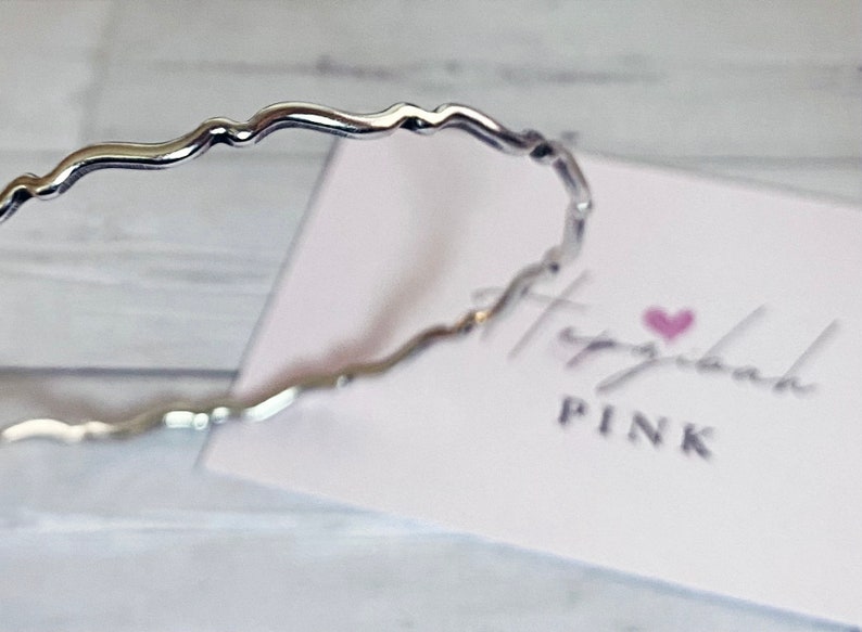 May include: A silver wavy bangle bracelet. The bracelet is on a white surface with a pink heart and the text "Hashtag Pink" on a white card.