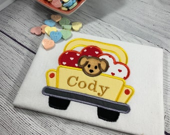 May include: A white fabric with an embroidered yellow truck carrying red and white hearts and a puppy. The name "Cody" is embroidered on the truck. A red heart-shaped bowl filled with candy hearts is in the background.