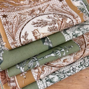 May include: A stack of folded fabric featuring a toile pattern in shades of ochre, olive green, and cream. The print displays detailed illustrations of winged figures, animals, birds, and botanical elements arranged within circular frames.