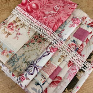 May include: A stack of folded fabric squares featuring various floral and botanical patterns in shades of pink, red, cream, and green. A strip of white decorative lace trim with a scalloped edge is draped diagonally across the top of the fabric.