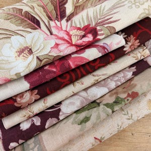 May include: A stack of folded fabric pieces featuring various floral patterns in shades of cream, beige, deep burgundy, and soft pink. The textiles display intricate botanical prints with large blossoms, leaves, and scrolling vines on a woven texture.