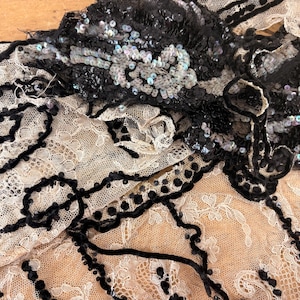May include: A piece of cream-coloured lace fabric featuring intricate black velvet ribbon trim and embellishments. The design includes dense clusters of shimmering black sequins and floral-inspired patterns, creating a textured contrast against the delicate, open-work lace base.