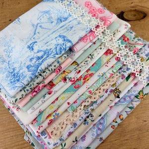 May include: A stack of assorted fabric squares featuring various floral, botanical, and toile patterns in shades of blue, pink, green, and yellow. The collection includes two strips of white and beige decorative lace trim layered across the top.