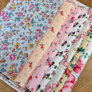 May include: A stack of folded fabric pieces featuring various floral patterns. The designs include a light blue background with pink and yellow flowers, a peach fabric with tiny red blossoms, and white fabrics with pink, red, and green floral prints.