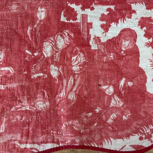 May include: A quilted fabric piece featuring a red and white toile de jouy pattern. The design depicts multiple figures in historical attire amidst foliage and trees. The textile has a diamond-shaped stitched pattern and a red fabric border.