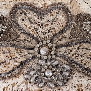 May include: An ornate lace appliqué featuring intricate embroidery, silver sequins, and white faux pearls. The design includes a central cluster of clear rhinestones and pear-shaped beads, surrounded by floral-shaped lace motifs and metallic thread.