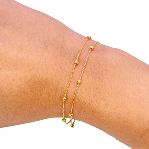 Könnte beinhalten: Ein zartes Goldarmband mit zwei feinen Ketten, die mit kleinen, gleichmäßig verteilten Perlen verziert sind. Das Armband wird an einem Handgelenk präsentiert und zeigt sein elegantes Design und seinen subtilen Glanz. Ein schlichtes, aber raffiniertes Schmuckstück.