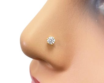 Puede incluir: Un piercing de nariz dorado con una circonita cúbica transparente y redonda, engastada con seis garras. El piercing está engastado en un metal color oro y se muestra en la nariz de un maniquí.