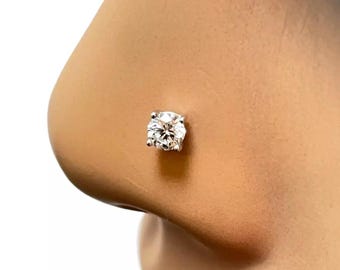 May include: A close-up of a nose with a single, clear, round-cut diamond nose stud. The diamond is set in a silver or white metal setting, and the skin tone is light.
