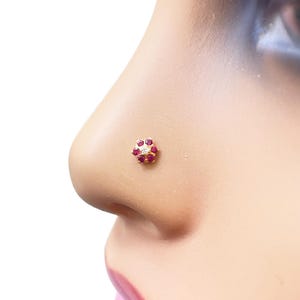 Puede incluir: Un piercing de nariz dorado con un diseño floral. Presenta seis pequeñas piedras preciosas rojas redondas que rodean una piedra preciosa central transparente. La joya está colocada en el lateral de la nariz de una persona.