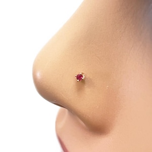 Pode incluir: Um piercing de nariz dourado com uma pequena pedra preciosa redonda de cor vermelha brilhante. O pino é colocado em uma delicada moldura dourada, criando uma joia simples, mas elegante. A joia é projetada para piercings no nariz.