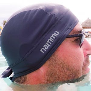 May include: A dark blue swim cap with the word "nammu" printed in white on the side. The cap is worn by a person in the water, with sunglasses. The background shows a beach scene with a thatched-roof structure.
