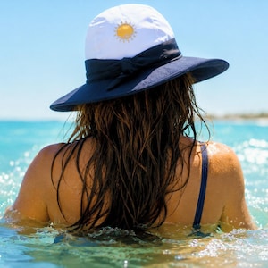 Puede incluir: Un sombrero de sol blanco con ala ancha de color azul marino y una cinta azul marino atada en un lazo en la parte delantera. La copa del sombrero presenta un pequeño icono de sol amarillo bordado en el centro del panel frontal.
