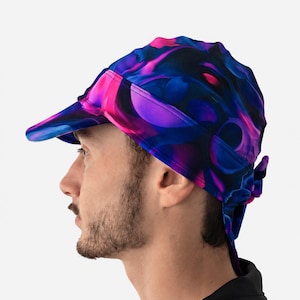 Puede incluir: Un gorro tipo skull cap con un patrón abstracto vibrante en tonos azul oscuro, morado y rosa brillante. La prenda incluye una visera corta y rígida en la parte delantera y lazos de tela en la parte posterior para un ajuste seguro y colorido.