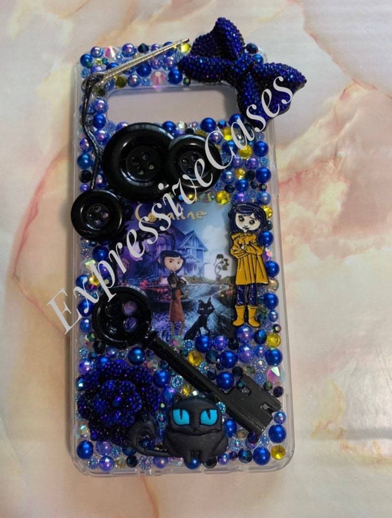 Puede incluir: Una funda de tel&eacute;fono transparente con un dise&ntilde;o azul y negro. La funda presenta una llave negra, un gato negro, una flor azul y un bot&oacute;n negro. La funda tambi&eacute;n presenta una imagen de una ni&ntilde;a con un impermeable amarillo y un gato negro. La funda est&aacute; decorada con diamantes de imitaci&oacute;n azules y negros.