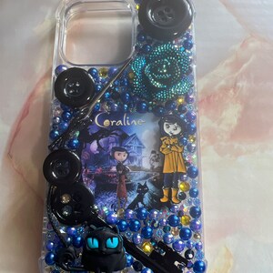 Puede incluir: Una funda transparente para tel&eacute;fono con un dise&ntilde;o negro y azul. La funda presenta una escena de la pel&iacute;cula Coraline, con el personaje principal, Coraline, y la Otra Madre. La funda est&aacute; decorada con botones negros, cuentas azules y amarillas, y un amuleto de gato negro.