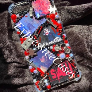 May include: Clear phone case decorated with red, black, and white beads, and small metal charms. The case features images and text from the movie "SAW". The text "Expressive Cases" is also visible.
