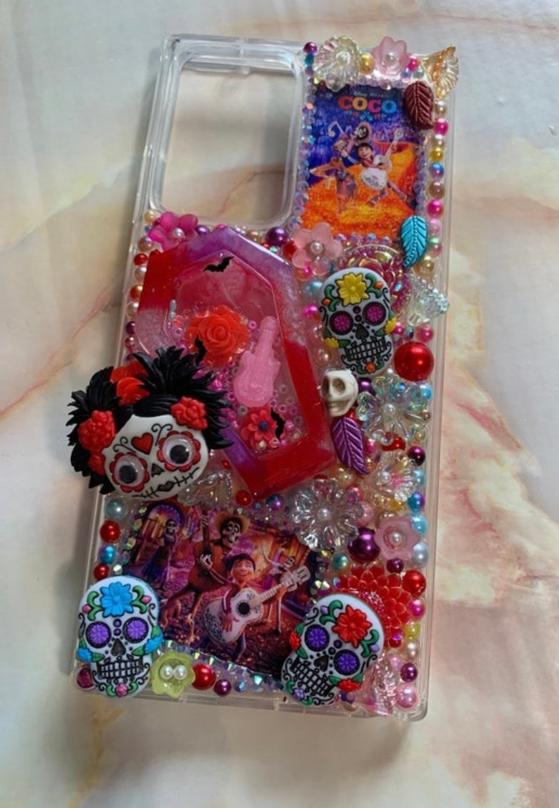 Puede incluir: Una funda transparente para tel&eacute;fono decorada con cuentas de colores, purpurina y abalorios. La funda presenta una imagen de los personajes de la pel&iacute;cula "Coco" y varios abalorios de calavera de az&uacute;car.