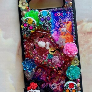 Puede incluir: Una funda de tel&eacute;fono negra decorada con calaveras de az&uacute;car de colores, flores y purpurina. La funda presenta una escena de la pel&iacute;cula de Disney Pixar "Coco".