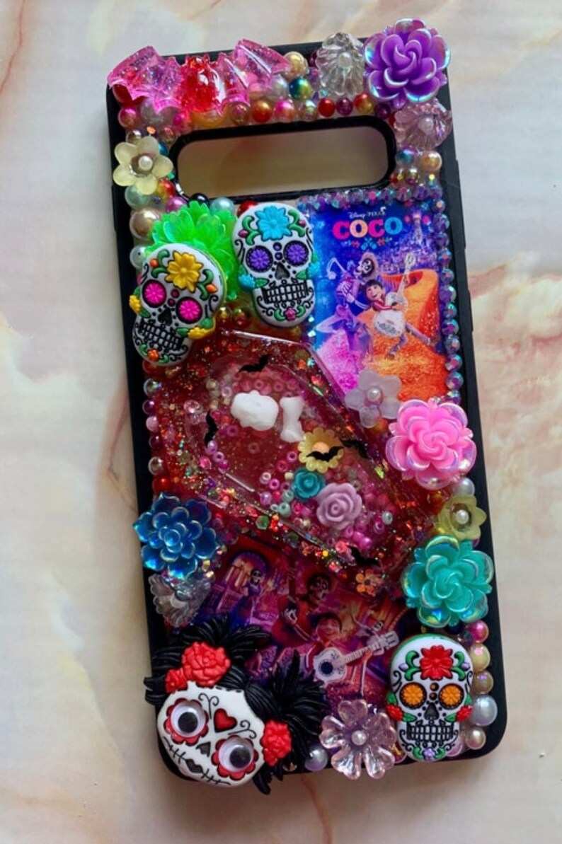 Puede incluir: Una funda de tel&eacute;fono negra decorada con calaveras de az&uacute;car de colores, flores y purpurina. La funda presenta una escena de la pel&iacute;cula de Disney Pixar "Coco".