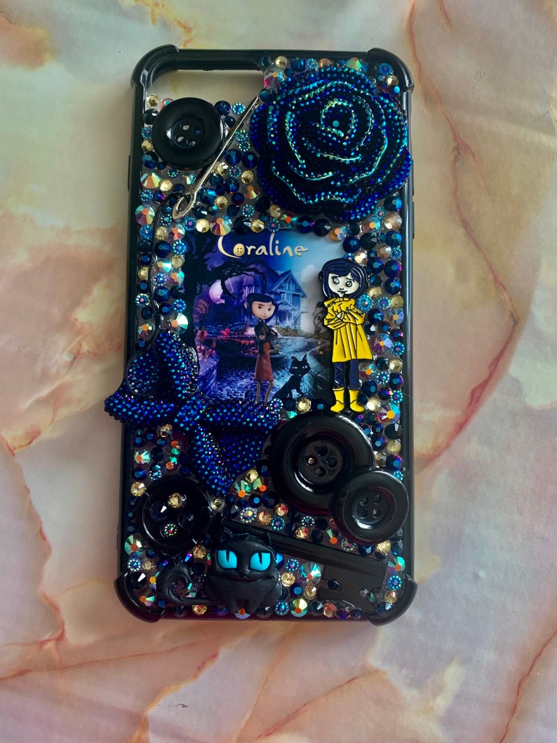 Puede incluir: Carcasa negra para iPhone con una rosa azul y negra, un gato negro, botones y una imagen de Coraline de la pel&iacute;cula Coraline.