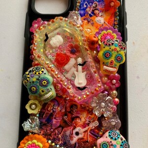 Puede incluir: Carcasa negra para iPhone decorada con calaveras de az&uacute;car de colores, flores y pedrer&iacute;a. La carcasa presenta un p&oacute;ster de la pel&iacute;cula 'Coco' y una secci&oacute;n de resina transparente con una guitarra blanca, huesos y un murci&eacute;lago.