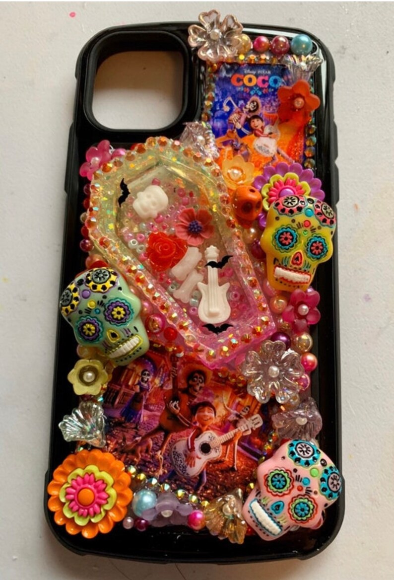 Puede incluir: Carcasa negra para iPhone decorada con calaveras de az&uacute;car de colores, flores y pedrer&iacute;a. La carcasa presenta un p&oacute;ster de la pel&iacute;cula 'Coco' y una secci&oacute;n de resina transparente con una guitarra blanca, huesos y un murci&eacute;lago.