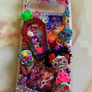 Puede incluir: Una funda transparente para tel&eacute;fono decorada con brillantes de colores, flores y calaveras de az&uacute;car. La funda presenta una imagen de los personajes de la pel&iacute;cula "Coco" y la palabra "Coco".