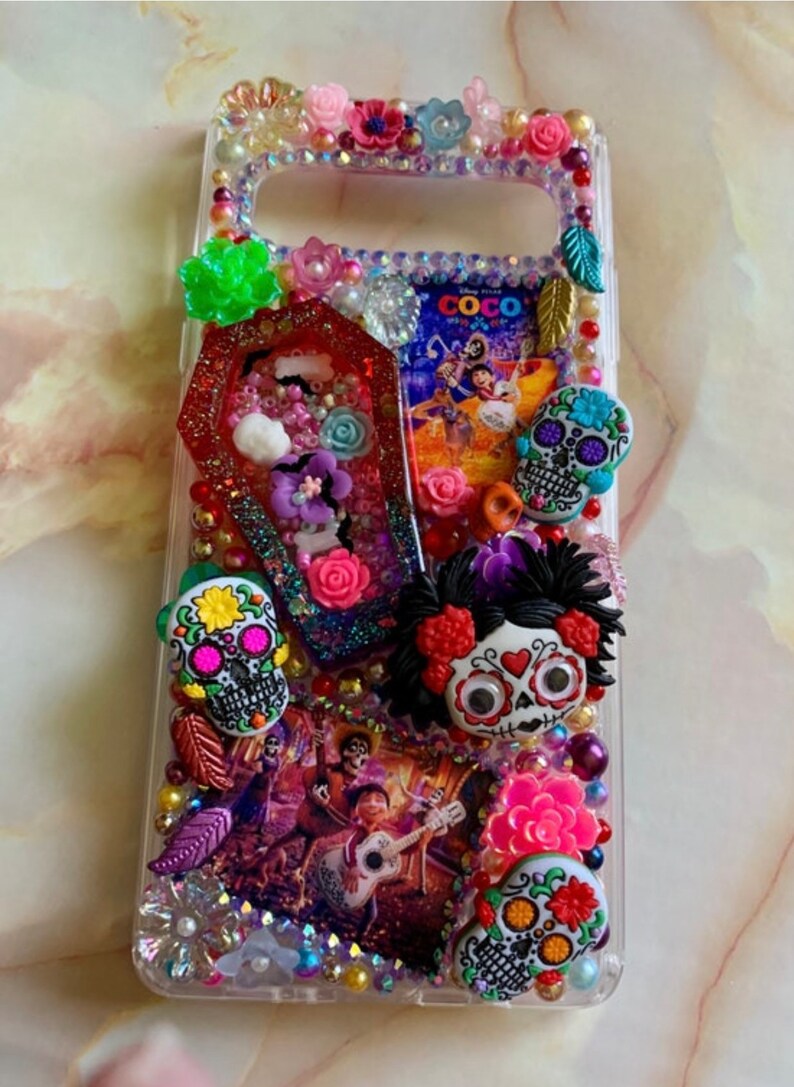 Puede incluir: Una funda transparente para tel&eacute;fono decorada con brillantes de colores, flores y calaveras de az&uacute;car. La funda presenta una imagen de los personajes de la pel&iacute;cula "Coco" y la palabra "Coco".