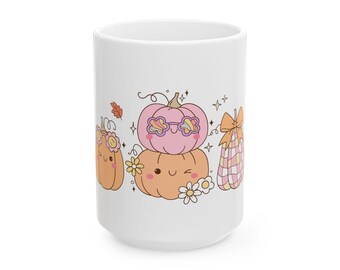 Op de afbeelding: Een witte keramische mok met een kleurrijke afbeelding van vier cartoonpompoenen met lachende gezichten. De pompoenen zijn versierd met bloemen, een ruitpatroon en steraccenten. Het ontwerp bevat zachte pastelkleuren zoals roze, oranje en geel.