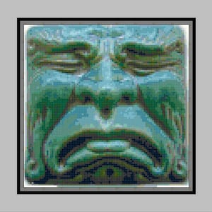 Könnte beinhalten: Nahaufnahme einer grünen Steinskulptur eines Gesichts mit einem traurigen Ausdruck. Das Gesicht hat tiefliegende Augen, einen breiten Mund und eine runzelige Stirn.