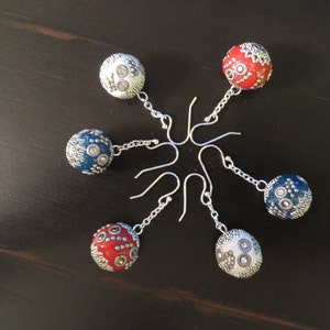 May include: Six pairs of dangle earrings with silver hooks and round, colorful, patterned beads. The beads are white, red, and blue with silver accents.