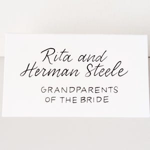 May include: A white card with black script that reads "Rita and Herman Steele" above "GRANDPARENTS OF THE BRIDE". The card is rectangular and sits on a white surface. The text is in a cursive font.