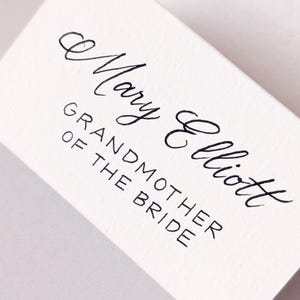 May include: A white card with the name "Mary Elliott" in elegant cursive script. Below, the text reads "Grandmother of the Bride" in a simple, sans-serif font. The card is angled, suggesting a formal event.