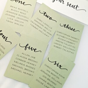 May include: Six green cards with the numbers one through six written in black cursive. Each card lists names in black text. The cards are on a white surface with the words "find your seat" written in black cursive.
