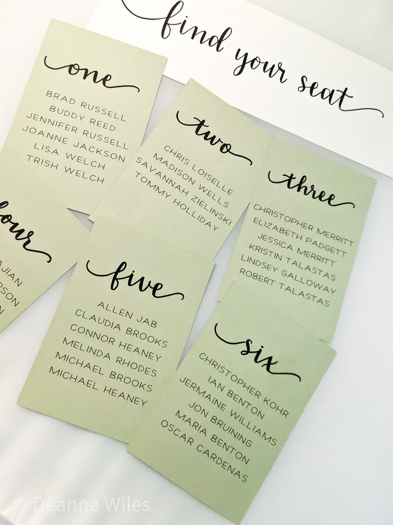 May include: Six green cards with the numbers one through six written in black cursive. Each card lists names in black text. The cards are on a white surface with the words "find your seat" written in black cursive.