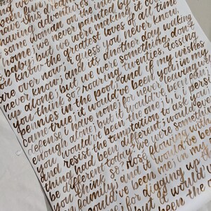 May include: A white sheet of paper with handwritten text in a metallic gold color. The text is a poem or a quote, with a cursive font style. The paper is slightly wrinkled, suggesting it is handmade.