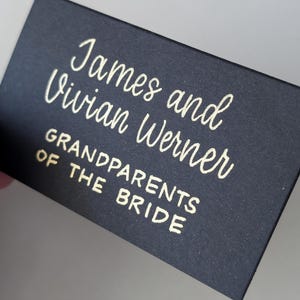 May include: A black place card with gold lettering. The card reads "James and Vivian Werner, GRANDPARENTS OF THE BRIDE" in a stylish script font. The card is folded at the top, creating a tent-like shape.
