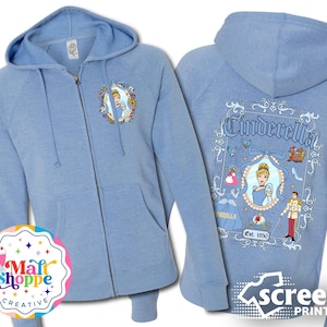 May include: Light blue zip-up hoodie with a screen printed design featuring Cinderella and Prince Charming. The design includes the text "Cinderella" and "Est. 1950".