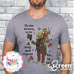 May include: A gray t-shirt with a Christmas-themed design featuring two green Muppet characters. The text on the shirt reads "Tis the Season to be Jolly and Joyous Fa la laa".