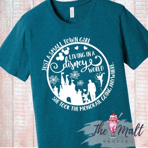 May include: Teal blue t-shirt with a white circle graphic. The graphic features a silhouette of Cinderella's castle, fireworks, a fairy, and a family. The text reads "Just a small town girl living in a disney world. She took the monorail going anywhere."
