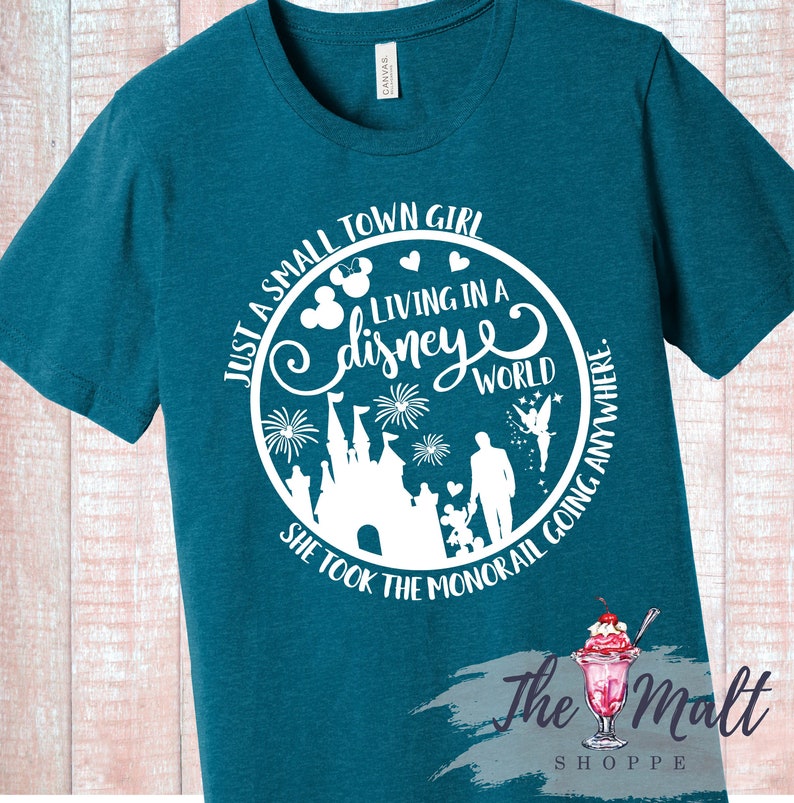 May include: Teal blue t-shirt with a white circle graphic. The graphic features a silhouette of Cinderella's castle, fireworks, a fairy, and a family. The text reads "Just a small town girl living in a disney world. She took the monorail going anywhere."