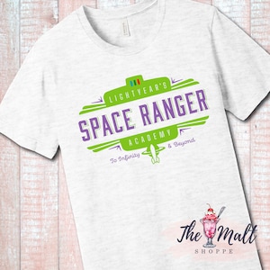 May include: White t-shirt with a green and purple logo that says "Lightyear's Space Ranger Academy To Infinity & Beyond". The logo is in a retro style with a spaceship graphic. The t-shirt is from The Malt Shoppe.