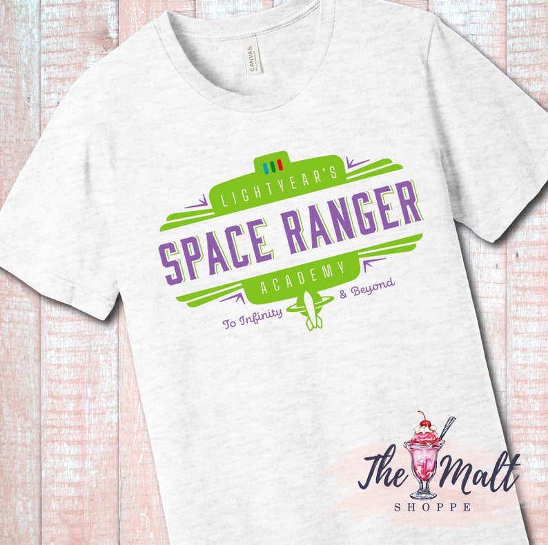May include: White t-shirt with a green and purple logo that says "Lightyear's Space Ranger Academy To Infinity & Beyond". The logo is in a retro style with a spaceship graphic. The t-shirt is from The Malt Shoppe.