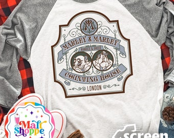 Op de afbeelding: Een wit en grijs baseballshirt met een bruin en wit grafisch ontwerp. De grafiek toont een logo in vintage stijl met de tekst "Marley & Marley Counting House London" en twee cartoonfiguren.