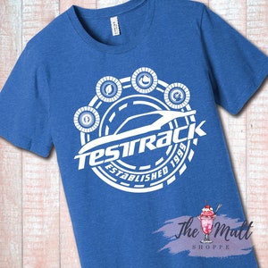 May include: A royal blue t-shirt with a white graphic design. The graphic features a stylized car with the text "Testrack Established 1999" around it. There are five small circles around the car with different symbols inside.